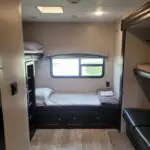 Compact RV bunk bedroom with a single bed along the wall, window, and built-in dark wood storage drawers underneath.