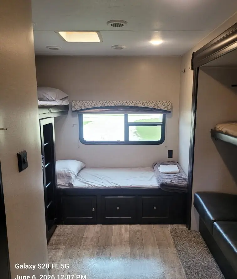 Compact RV bunk bedroom with a single bed along the wall, window, and built-in dark wood storage drawers underneath.