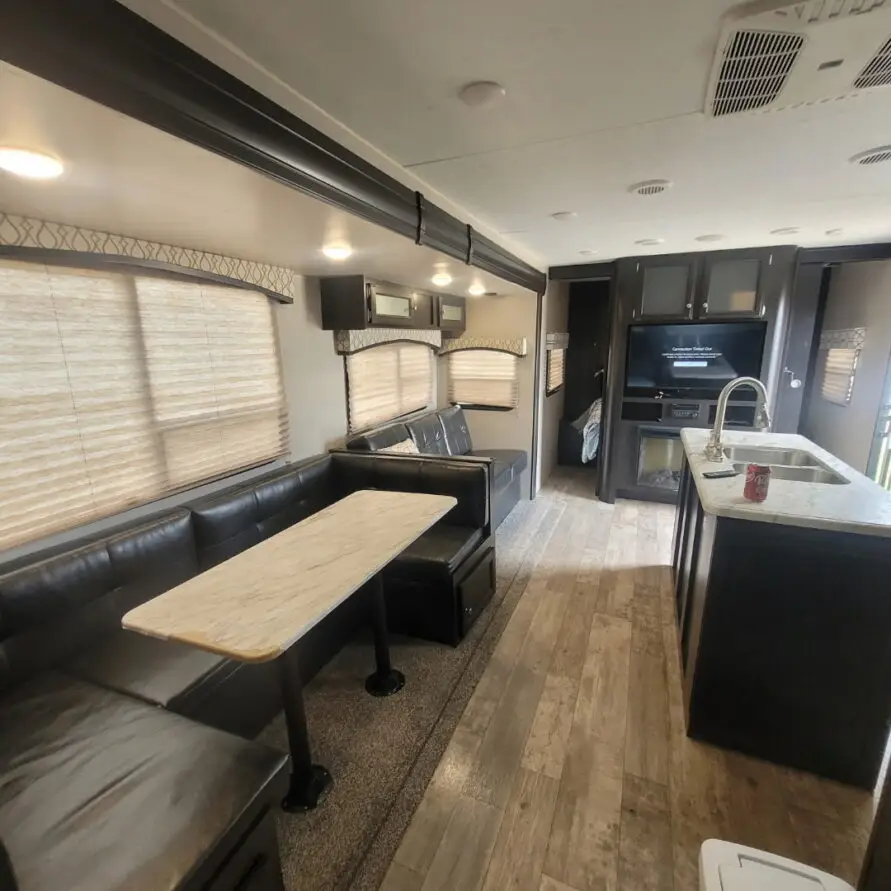 Interior of an RV living area with a black leather L-shaped sofa, marble dining table, and kitchen island with sink nearby.