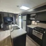 Interior view of a modern RV kitchen with dark cabinets, marble countertop island, double sink, and stainless steel appliances (stove, microwave, fridge).