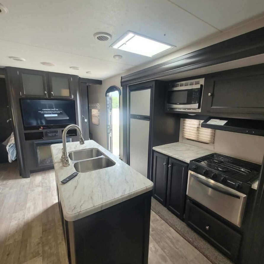 Interior view of a modern RV kitchen with dark cabinets, marble countertop island, double sink, and stainless steel appliances (stove, microwave, fridge).