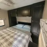 Small bedroom in an RV with a double bed, dark wood overhead cabinets, and a built-in headboard shelf above the mattress bedspread in plaid tones.