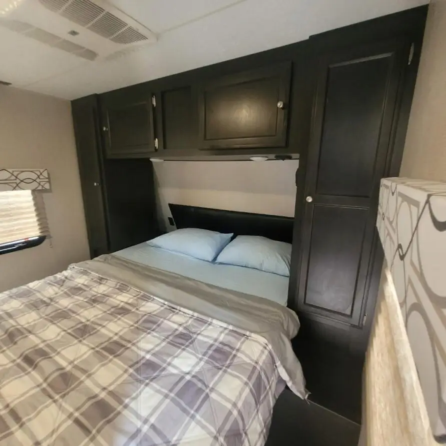 Small bedroom in an RV with a double bed, dark wood overhead cabinets, and a built-in headboard shelf above the mattress bedspread in plaid tones.
