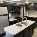 RV kitchen inside a travel trailer with a marble-look island sink, stainless steel faucet, stove and microwave, a side fridge, and dark cabinets