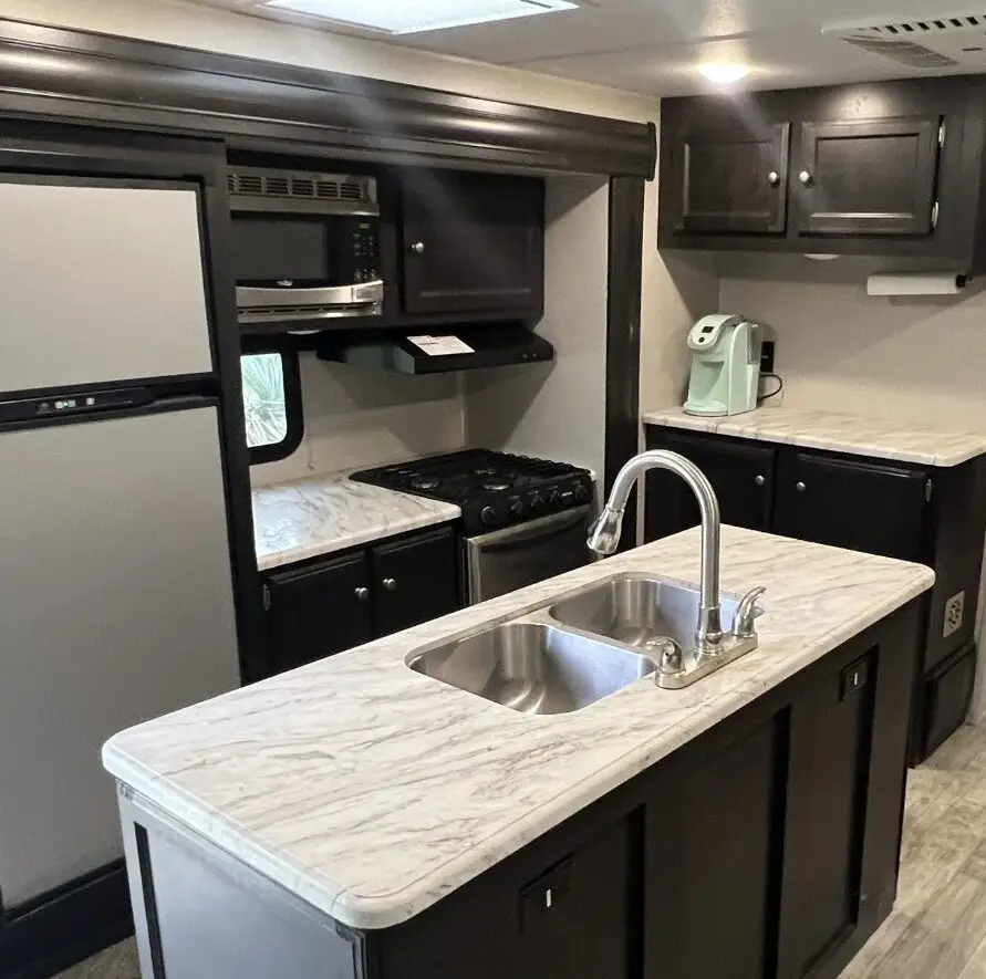 RV kitchen inside a travel trailer with a marble-look island sink, stainless steel faucet, stove and microwave, a side fridge, and dark cabinets