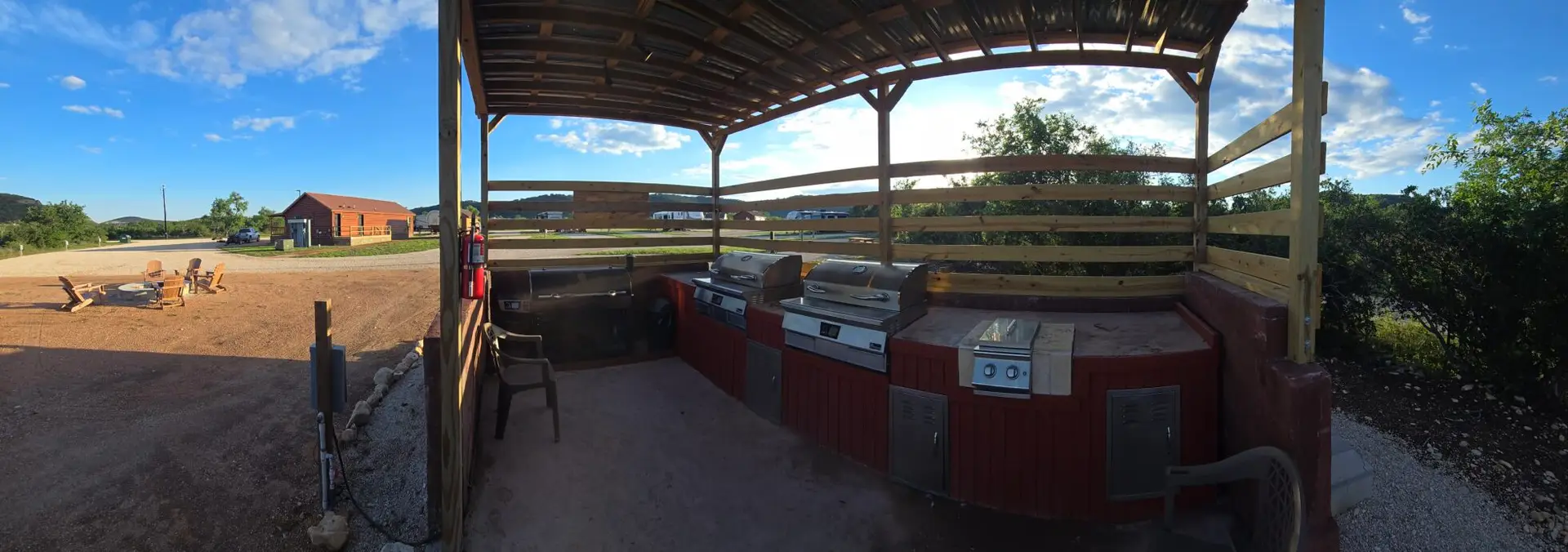 Outdoor covered grilling area with several stainless grills and a red wooden counter, set in a campground yard under a blue sky with trees nearby.