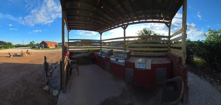 Outdoor covered grilling area with several stainless grills and a red wooden counter, set in a campground yard under a blue sky with trees nearby.