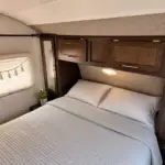 A cozy RV bedroom with a queen bed, white pillows, and a dark wood overhead cabinet; small nightstand with a plant beside the window.
