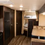 Interior of a compact RV: kitchen with microwave, oven, and fridge on the left; dining booth and window seating on the right; bunk beds toward the rear.
