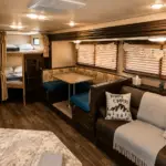 Interior of a warmly lit camper van with a sofa, dining booth, and bunk beds along the back wall under soft ceiling lights.