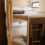 Compact bunk bed area inside a small camper: upper and lower twin beds with beige curtains and a small window nearby.